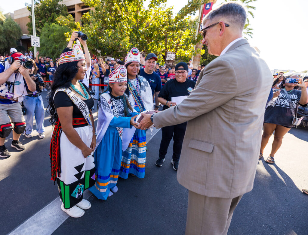 A person in a suit shakes hands with two individuals wearing traditional attire and ceremonial headdresses during an outdoor public event.
